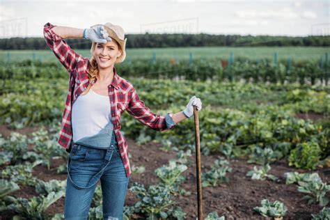 beautiful young female farmer in hat holding hoe and working on field ...